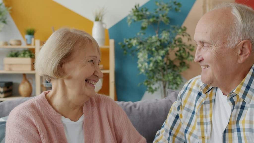An elderly couple smiling at each other on a couch.