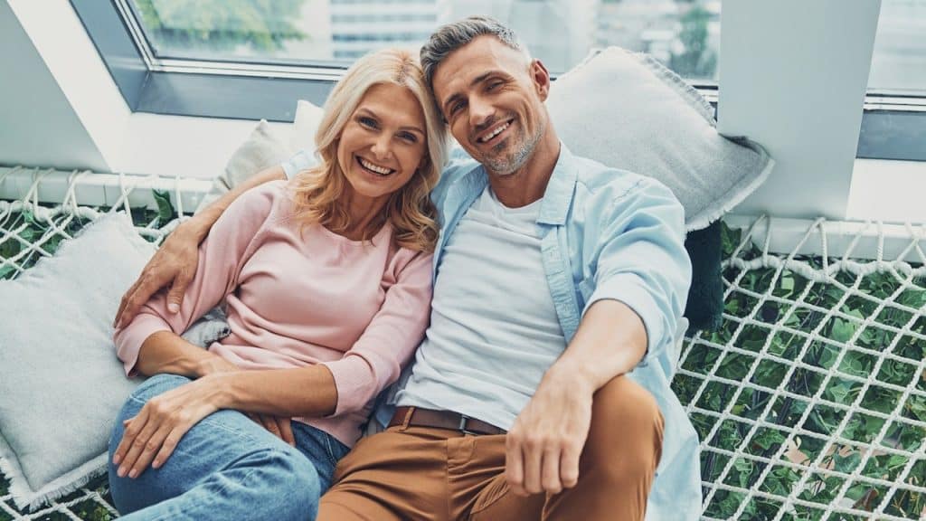 Happy mature couple smiling and looking at camera while relaxing in big hammock at home together.