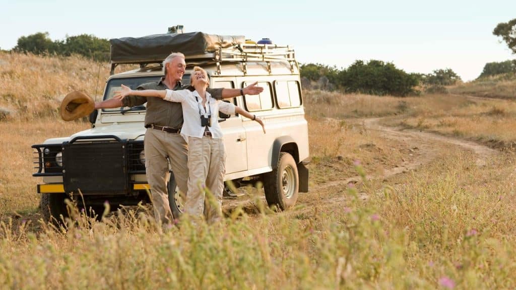 A happy, mature couple extending their arms in front of their jeep in a forest.