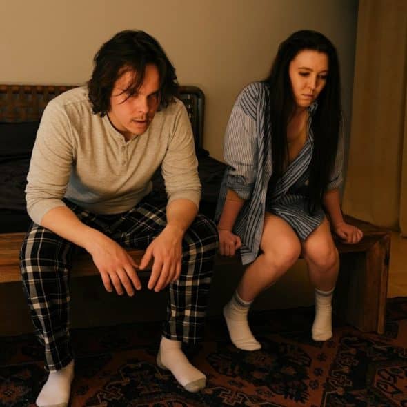 Two people sit on the edge of a bed in a dimly lit bedroom, both looking down and appearing tense or worried.