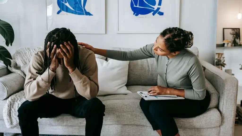 A woman sits on a couch, placing a hand on the shoulder of a distressed man.