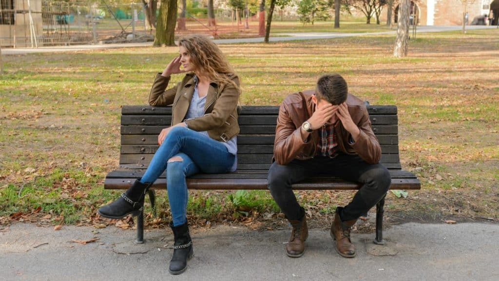 A man and woman sit on a park bench facing away from one another with heads down.