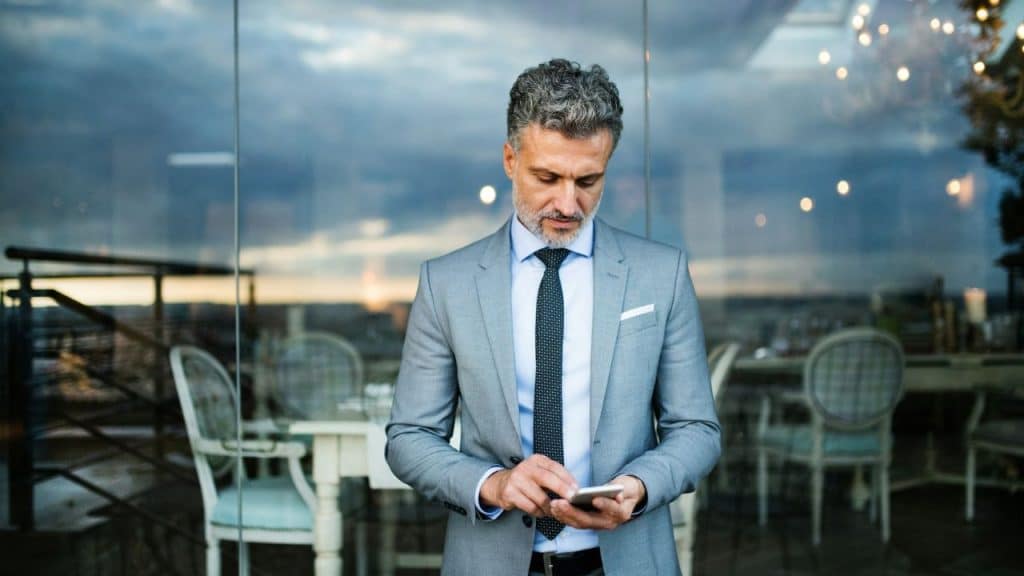 A man in a grey suit stands in front of a glass wall using a smartphone.