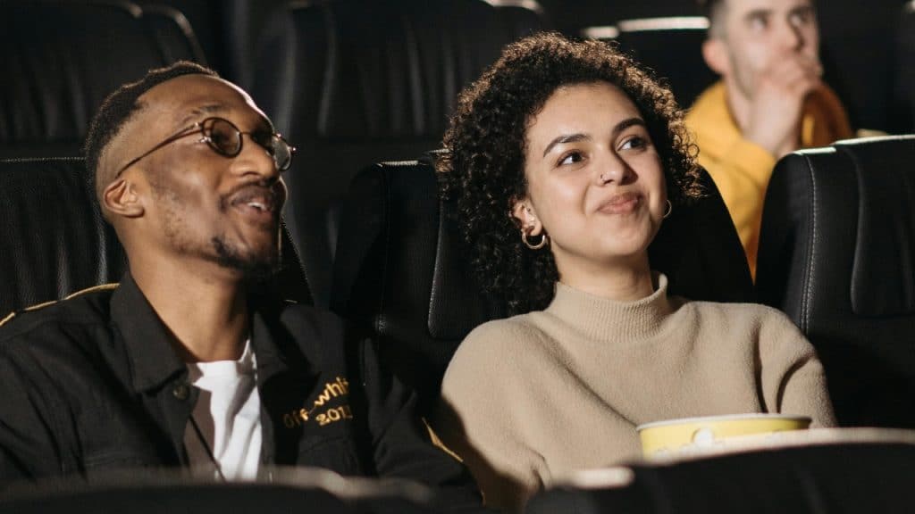Two people sit in dark theater seats, smiling and looking toward a screen with popcorn.