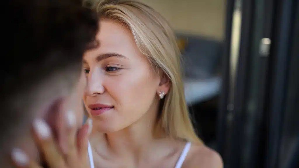 A close-up profile of a blonde woman smiling softly while a hand touches her face.