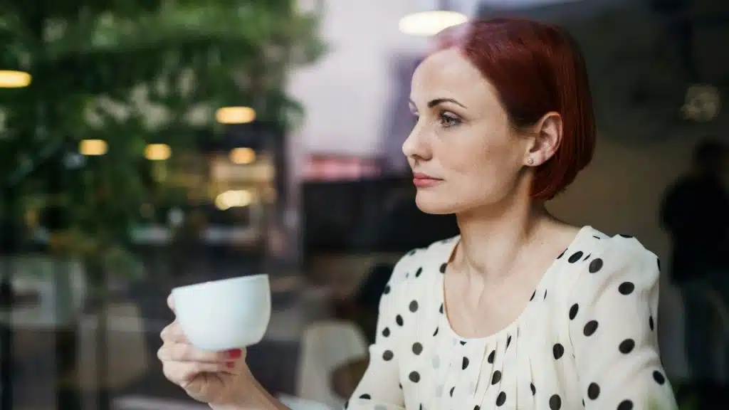 A woman with red hair holds a white cup while looking out of a window.