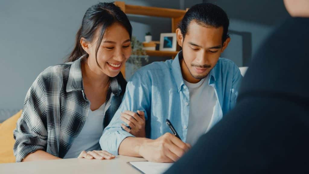 A smiling woman watches a man sign a document while sitting at a desk together.