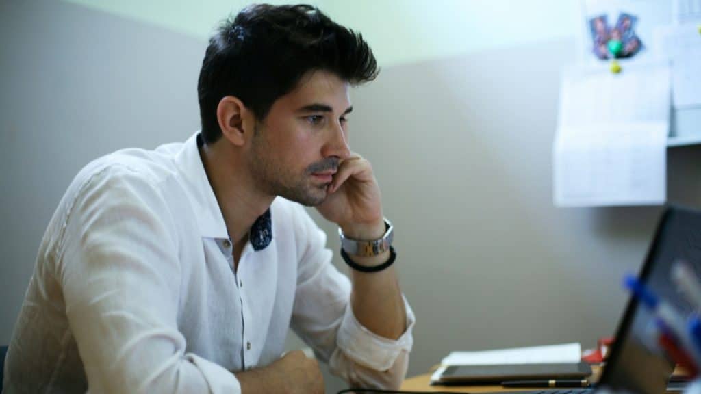 A man in a white shirt sits at a desk looking at a laptop computer screen.
