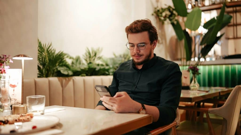 A man with glasses sits at a restaurant table alone, looking at his black smartphone.