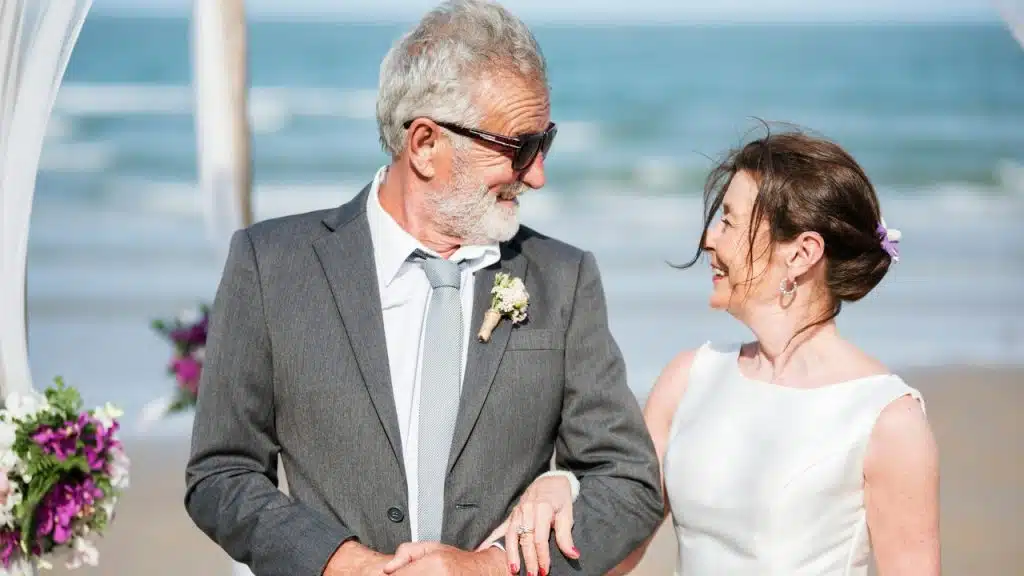 An older man in a grey suit and sunglasses smiles at a woman in white.