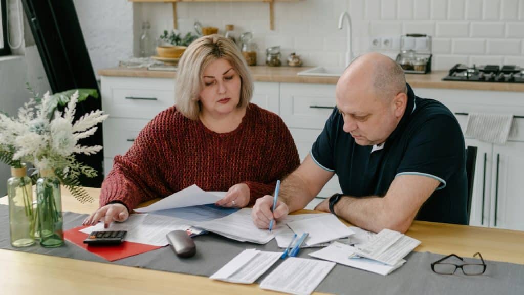 A man and woman sit at a kitchen table using a calculator and many papers.
