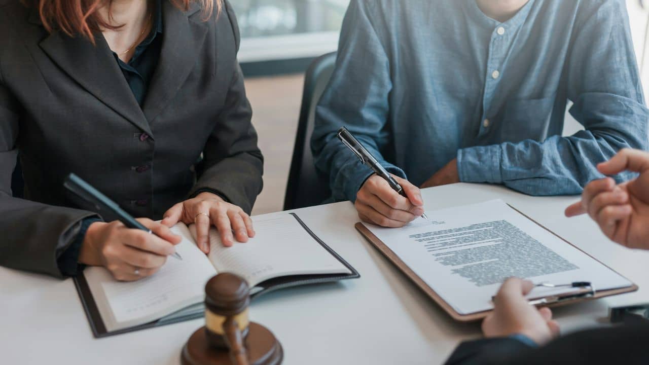 Two people sit at a desk signing and writing in documents next to a wooden gavel.