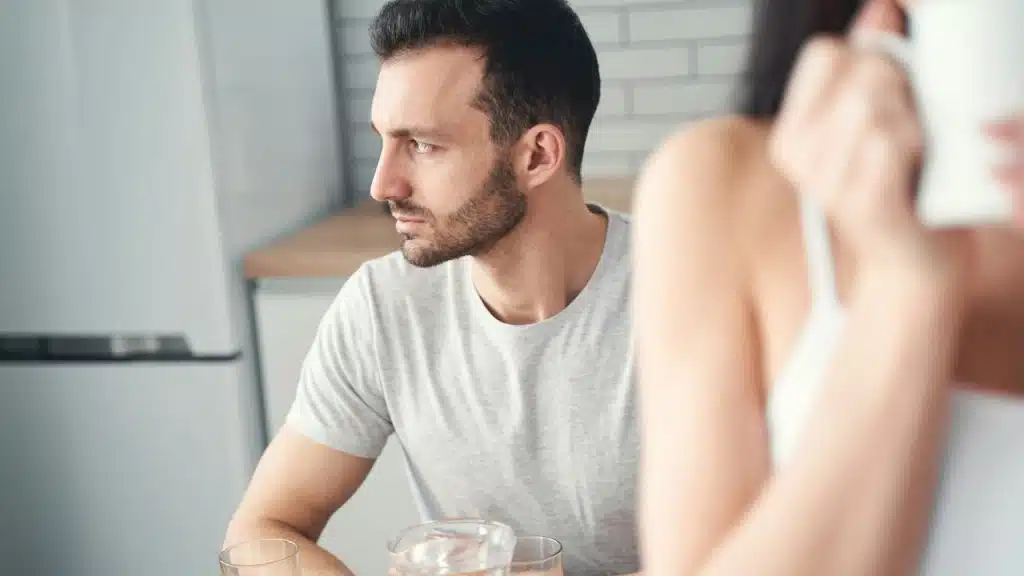 A woman having coffee is talking on the phone while her husband sitting behind her is looking on the side.