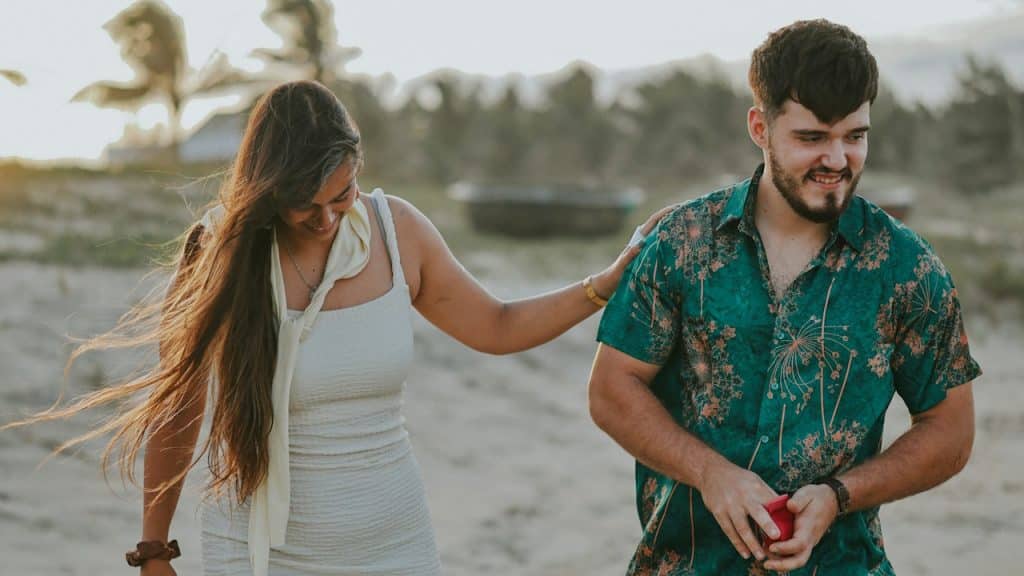 A couple walking on a beach, the man holding a ring box in his hands.