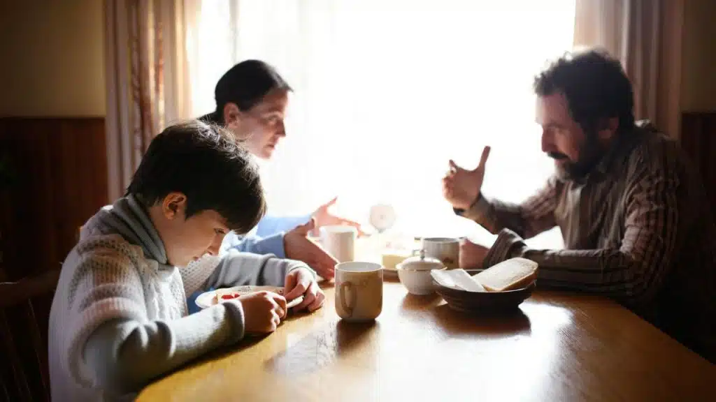 A child eats at a table while two adults talk in the background near a window.