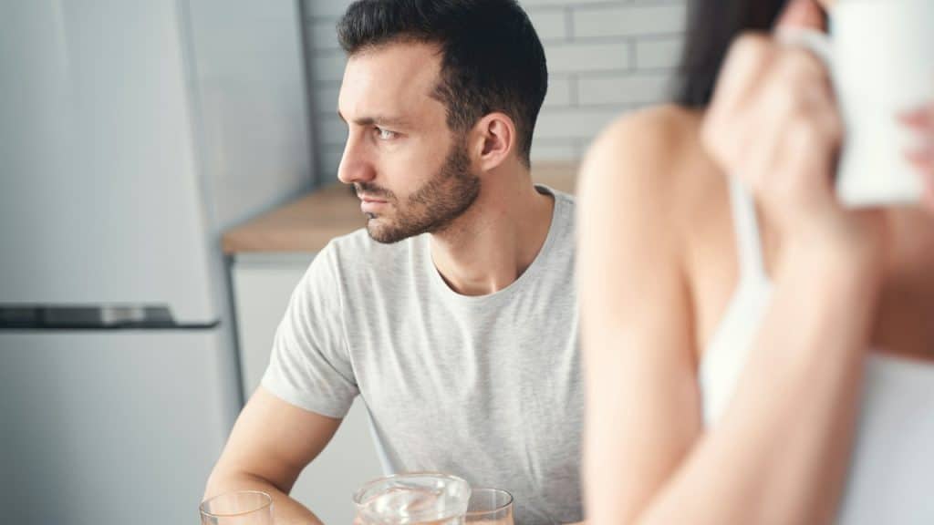 A blurred image of a woman holding a cup of coffee while a man is sitting behind her, quietly looking away.