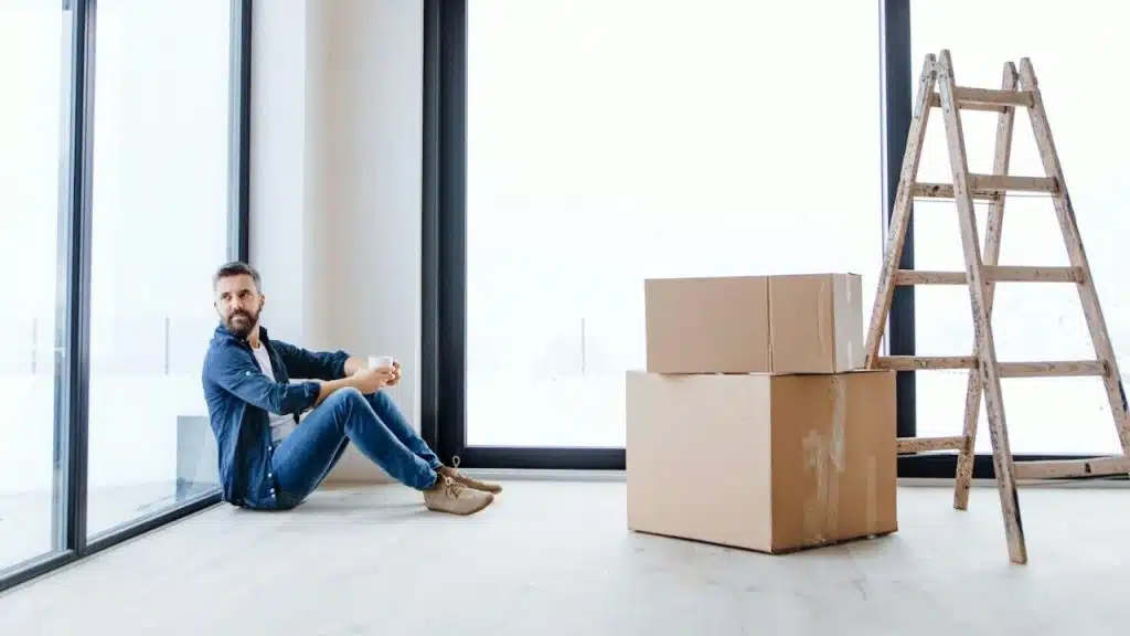A man sits on the floor near cardboard boxes and a wooden ladder by windows.