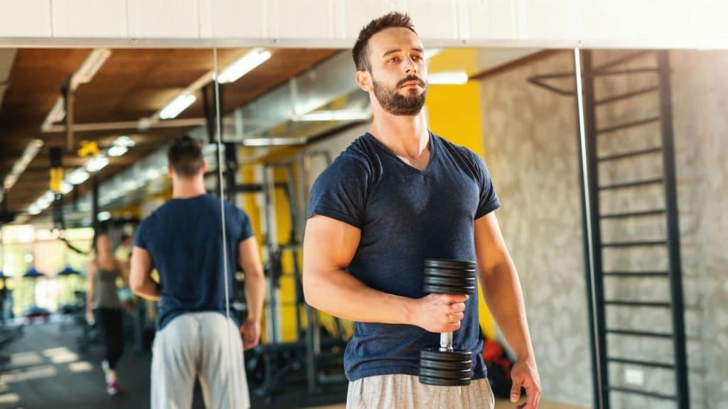 A man in a blue t-shirt stands in a gym, holding a black dumbbell.