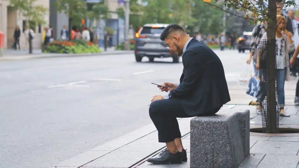 A man in a suit sits on a stone block outdoors looking at a mobile phone.