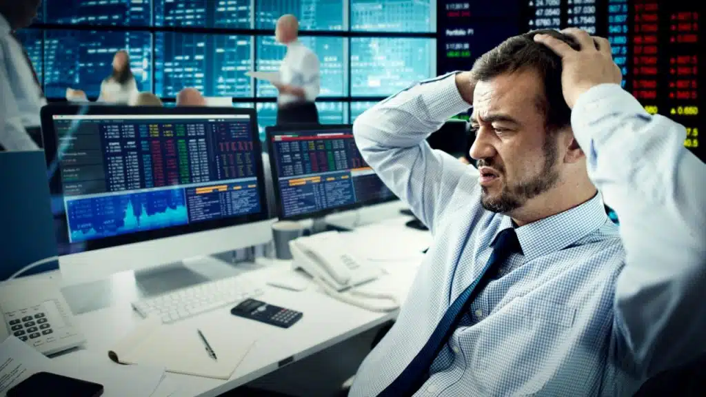 A man in a shirt and tie holds his head while looking at computer monitors.