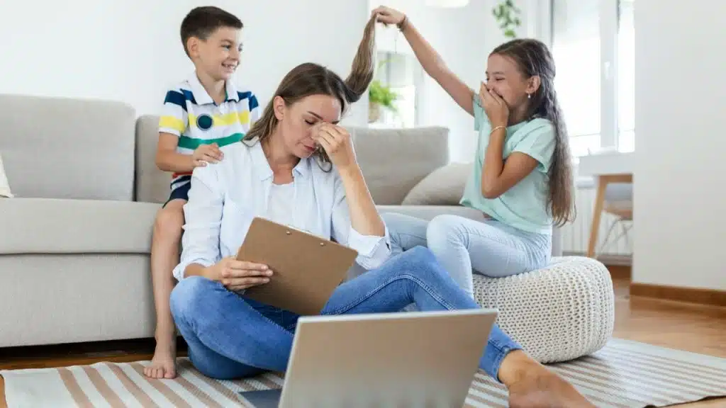 A woman sits on the floor with a clipboard while two children play with her hair.