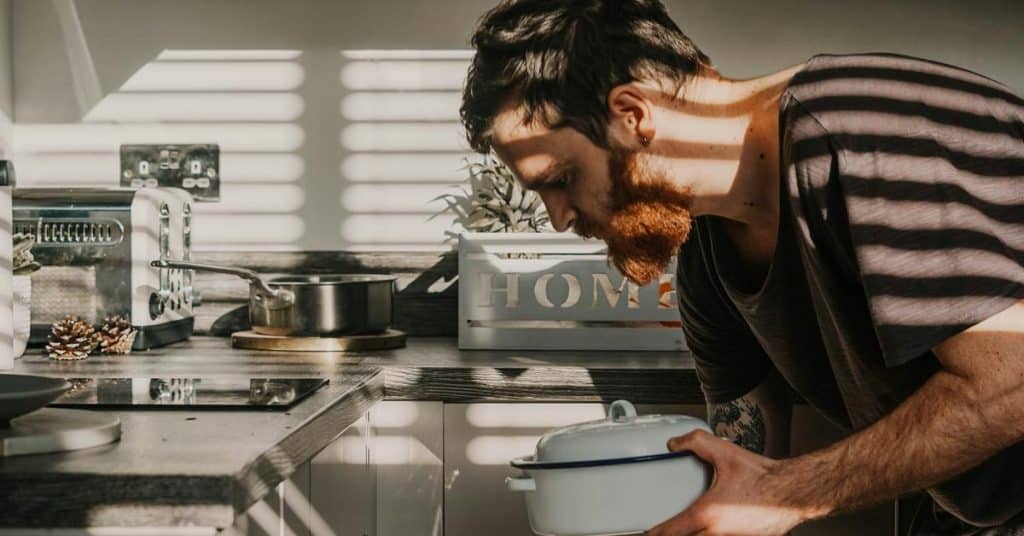 A bearded man cooking in a sunlit kitchen while holding a pot.