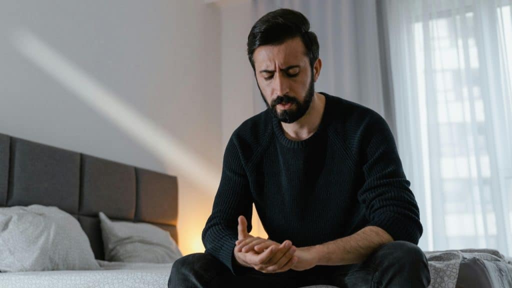 A bearded man in a dark sweater sits on a bed with his head bowed down.