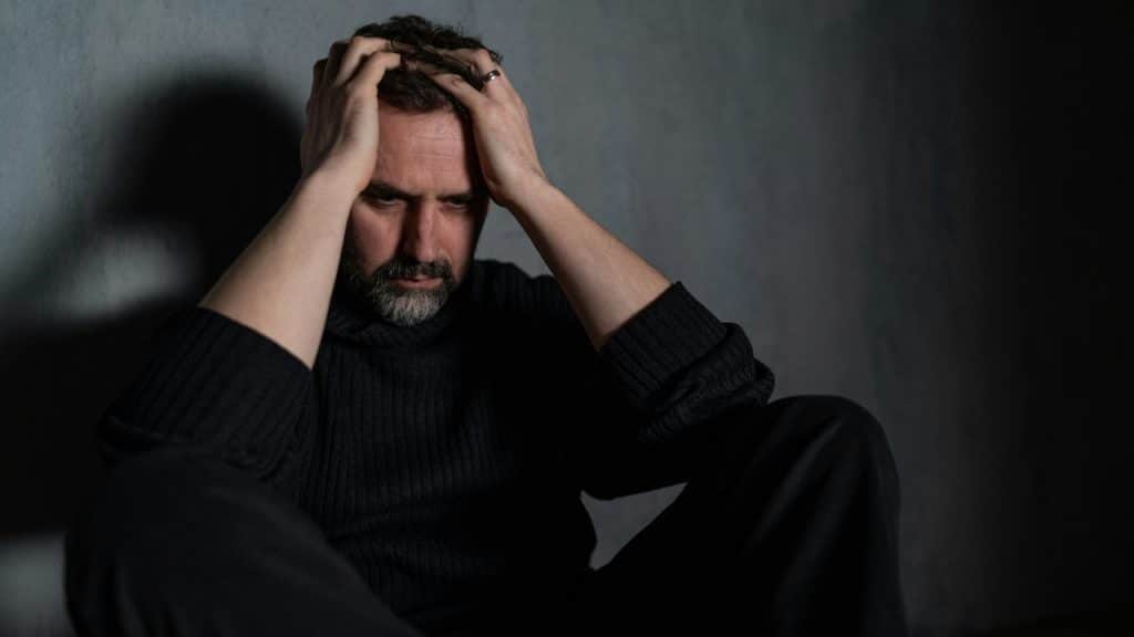 A bearded man in a dark sweater sits against a wall with hands in his hair.
