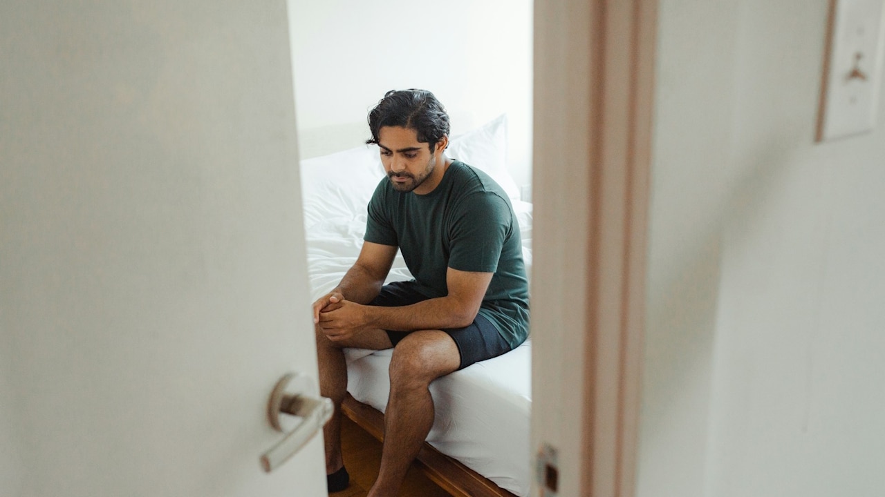 A lonely man sitting by himself on the edge of a bed in a doorway, looking down.