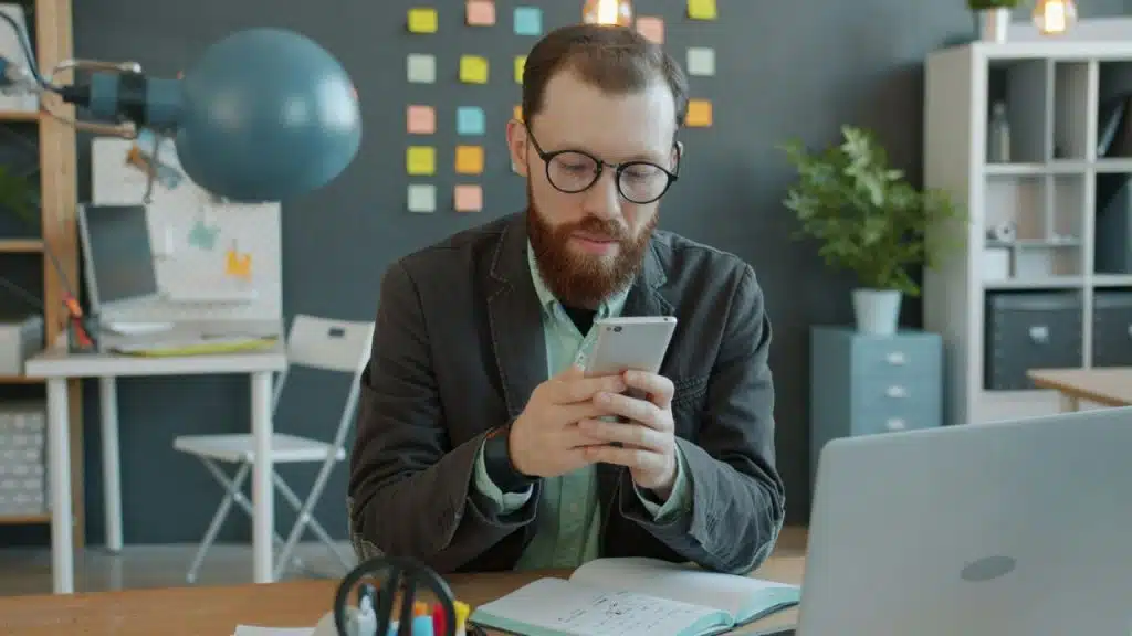 A bearded man with glasses uses a smartphone at a desk with a laptop and notebook.