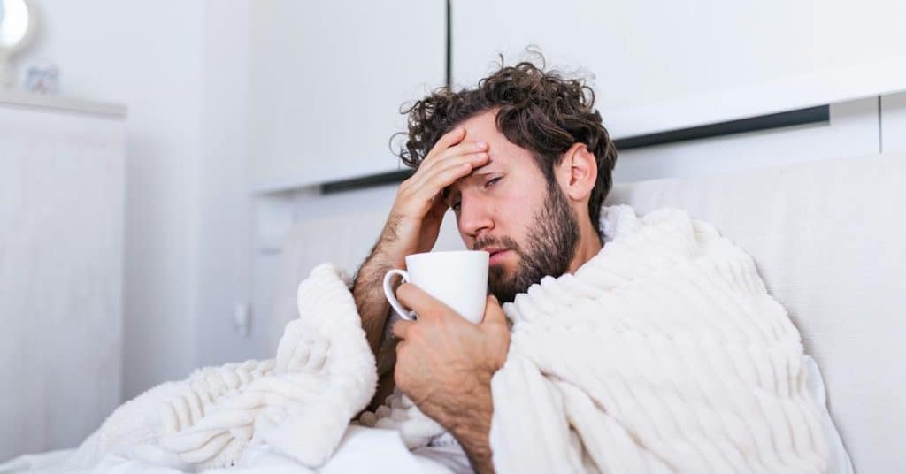 A man wrapped in a blanket holds a mug and rests his hand on his forehead.