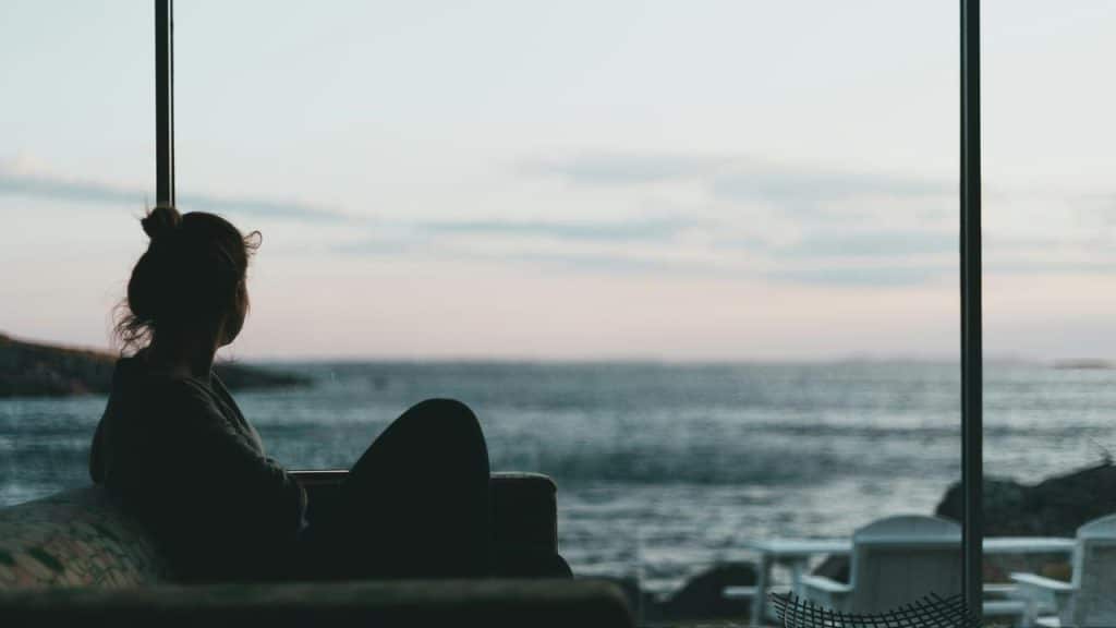A silhouetted person sitting by a large window, gazing out at the calm ocean.
