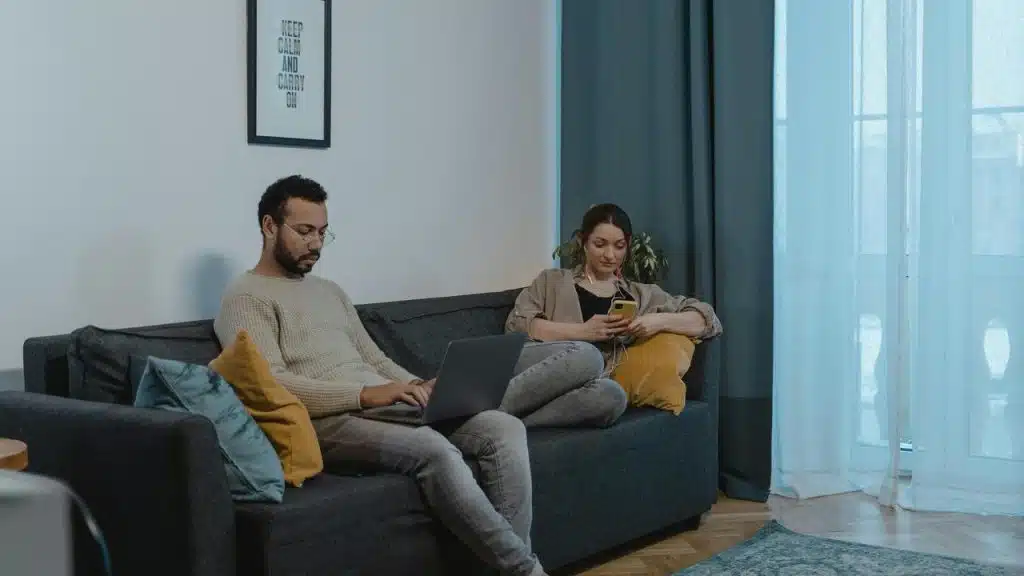 A couple sitting on a couch using a laptop and a phone.