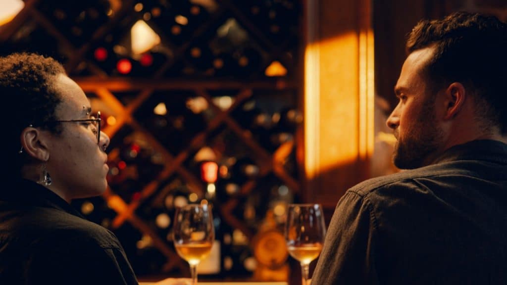 Two people sit at a bar with wine glasses in front of a wine rack.