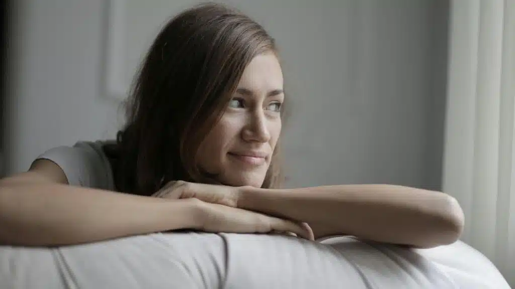 A woman resting her arms on a couch and looking out a window.