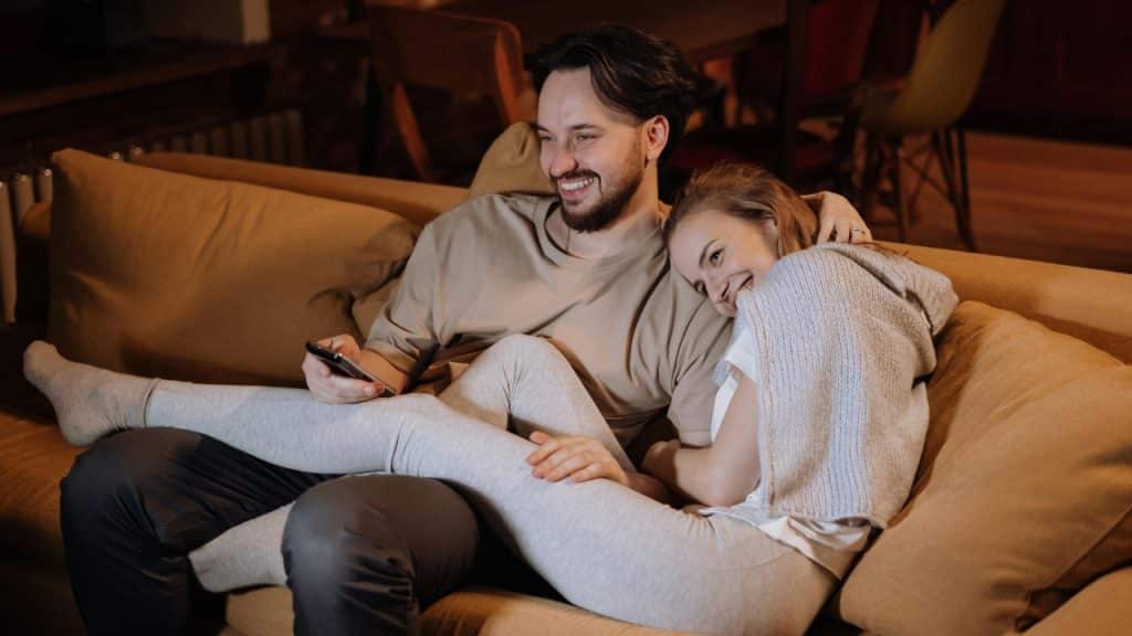 A man and woman sit closely on a couch, smiling while looking at a smartphone.