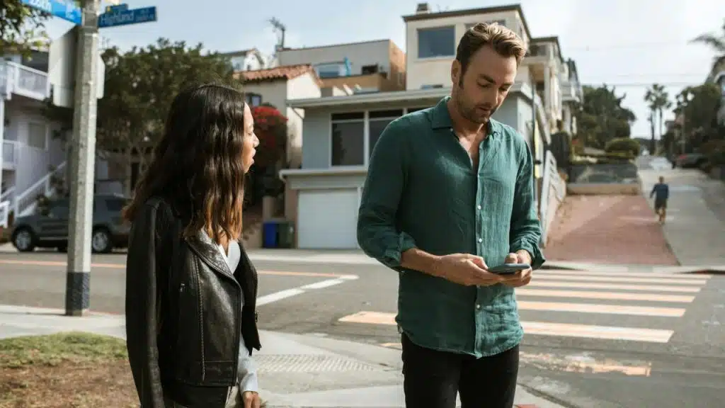A man looks at a phone while a woman in a leather jacket stands beside him.