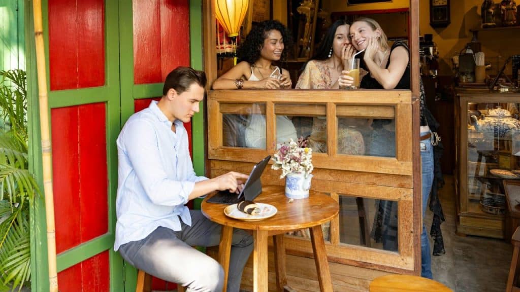 A man sits at an outdoor table with a tablet while three women whisper nearby.