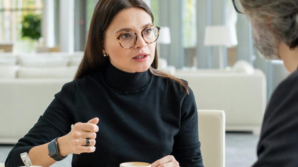 A woman wearing glasses gestures while talking to someone across a table.