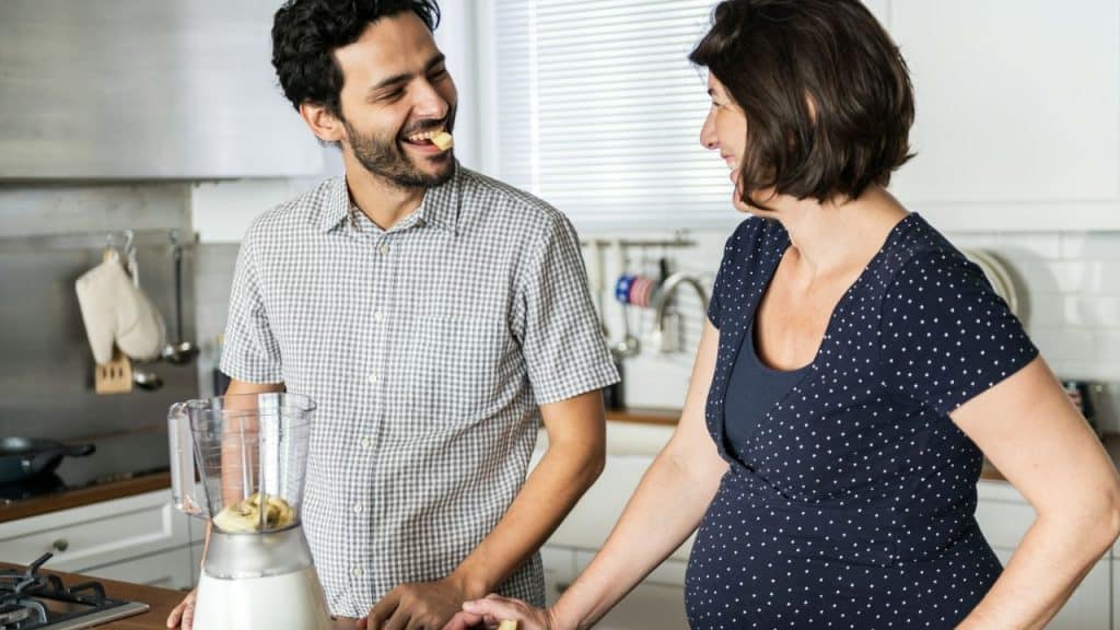 A laughing man with food in his mouth stands by a blender with a woman.