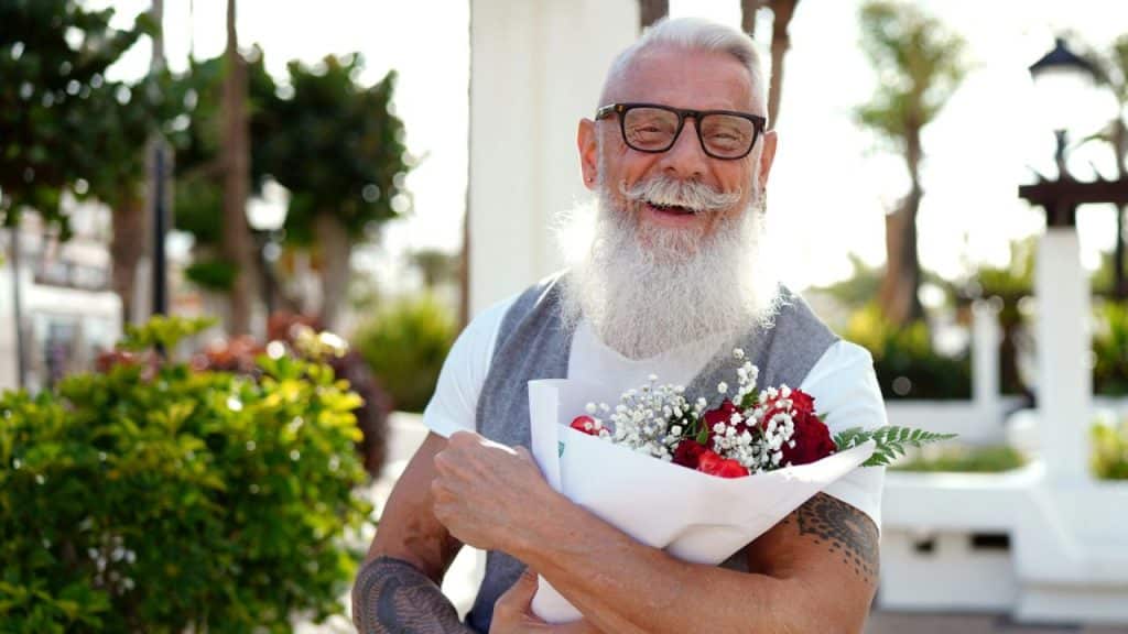 A smiling man with a long white beard and tattoos holds a bouquet of flowers.