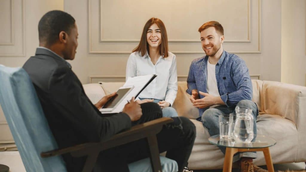 A man and woman sit on a couch smiling toward a man holding a clipboard.