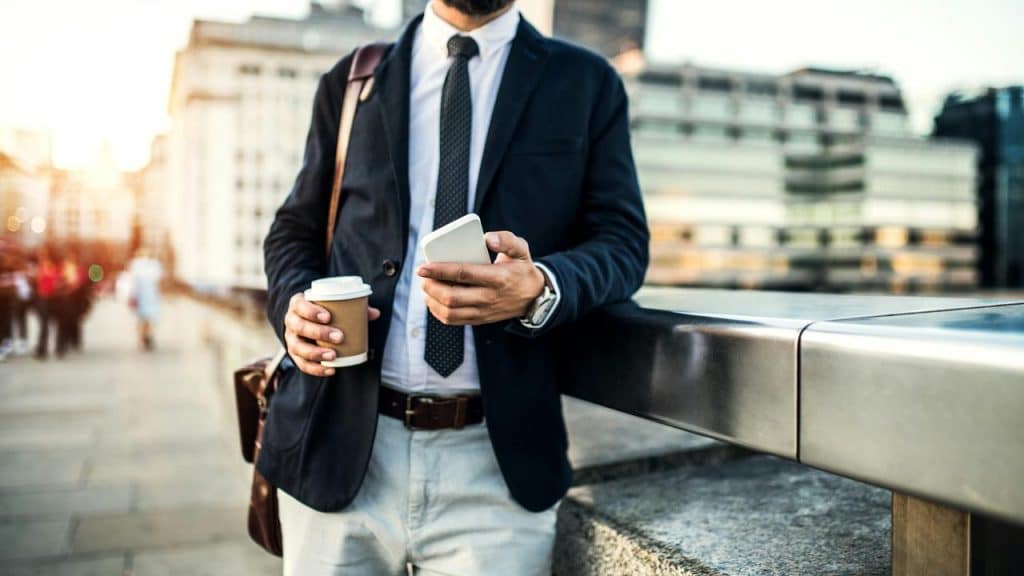 A businessman holding a coffee and checking his phone on a city walkway.