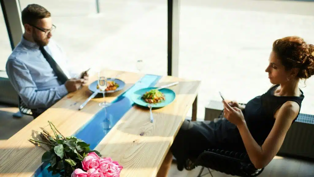 A man and woman sit at a dining table while both look at their smartphones.