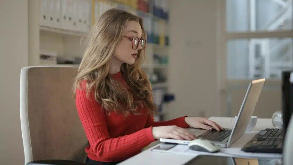 A blonde woman wearing red glasses and a red sweater types on a silver laptop.