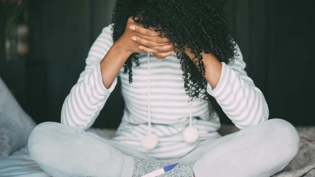 A woman sitting cross-legged on a bed, covering her face with her hands.