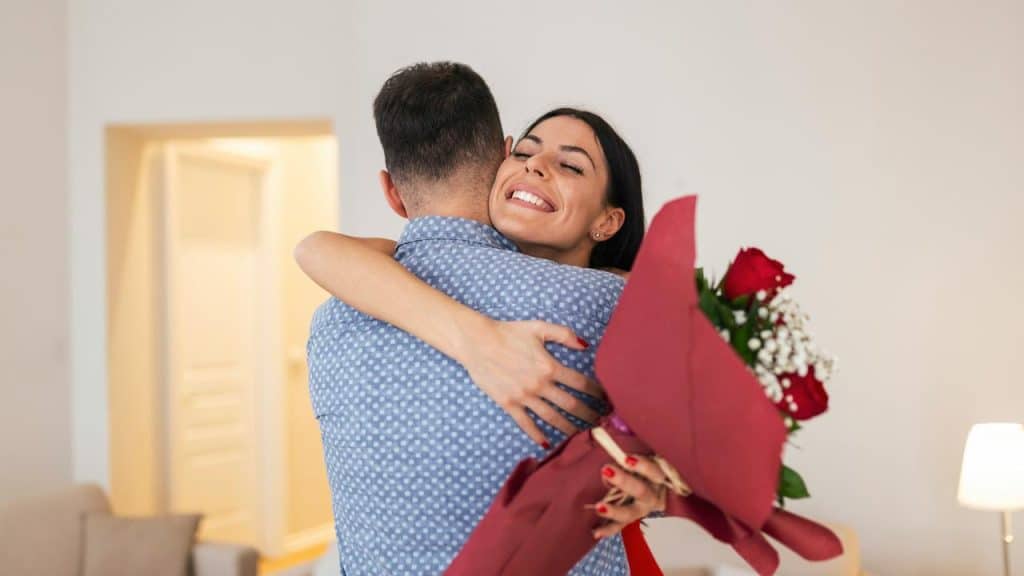 A smiling woman hugging a man while holding a bouquet of red roses.