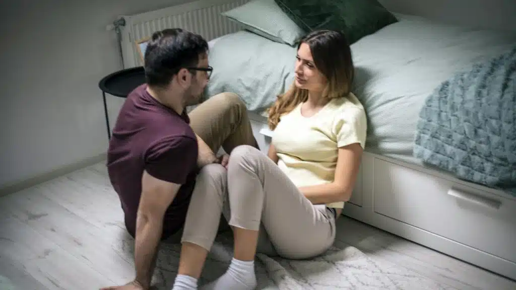 A couple sitting on the floor by a bed and talking quietly.