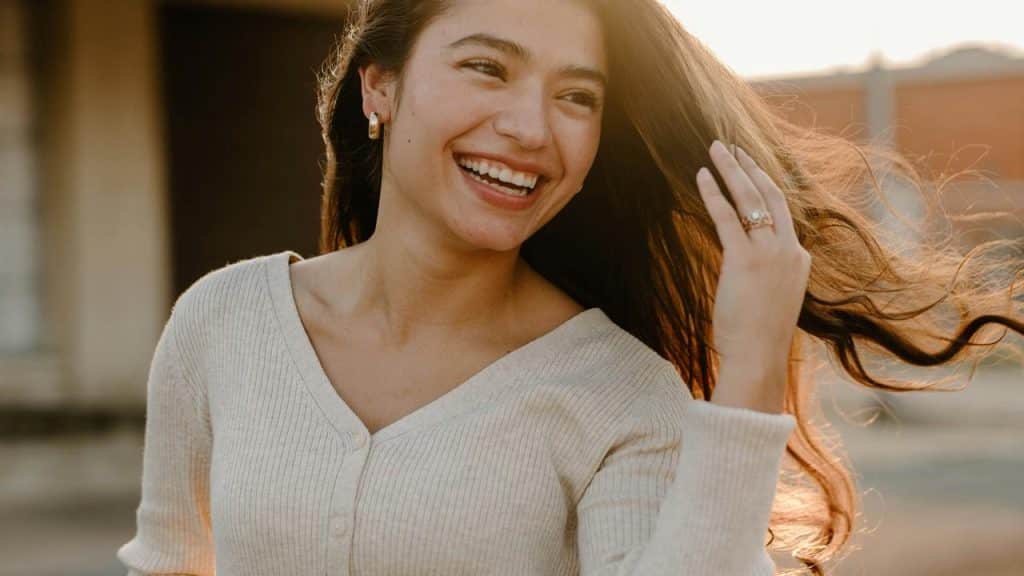 A smiling woman with long hair laughing outdoors in warm sunlight.