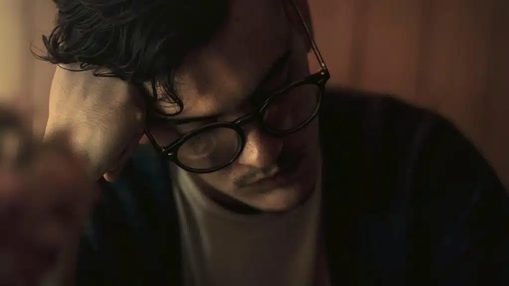 A close-up of a man wearing glasses, resting his head on his hand.
